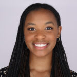 A person smiling with long black hair in braids, wearing a black shirt, in front of a light grey background.
