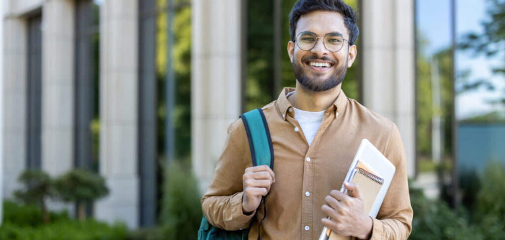 Smiling student stands outside academic building holding backpack and notebooks.