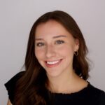 A person smiling with long dark brown hair in a dark shirt in front of a neutral background.