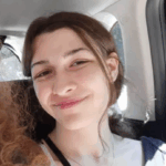 A person smiling with their dark hair pulled back, sitting a backseat of a car.