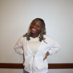 A person smiling with short dark hair in braids with their hands on their hips, in a light hoodie standing in front of a background.