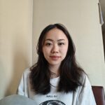 A person with long, dark brown hair and a neutral expression, wearing a white shirt in front of a neutral background.