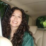 A person smiling with long, dark brown curly hair sitting in the backseat of a car with tan seats.