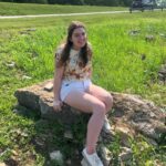 A person smiling with long light brown hair, wearing a light shirt and white shorts, sitting on a large rock in a field of green grass.
