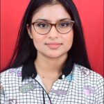 A person with long, straight, dark brown hair, wearing glasses and a patterned shirt, in front of a red background.