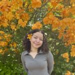 A person smiling with dark brown, short curly hair, wearing a grey shirt, with a yellow flower in their hair, standing front of a tree with yellow flowers.