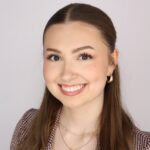 A person with long brown hair, smiling in front of a light grey background.