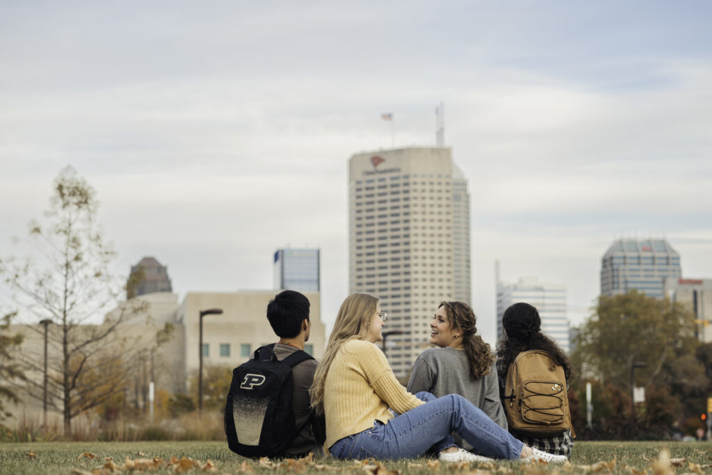 Three students sitting on the grass facing Indy buildings.