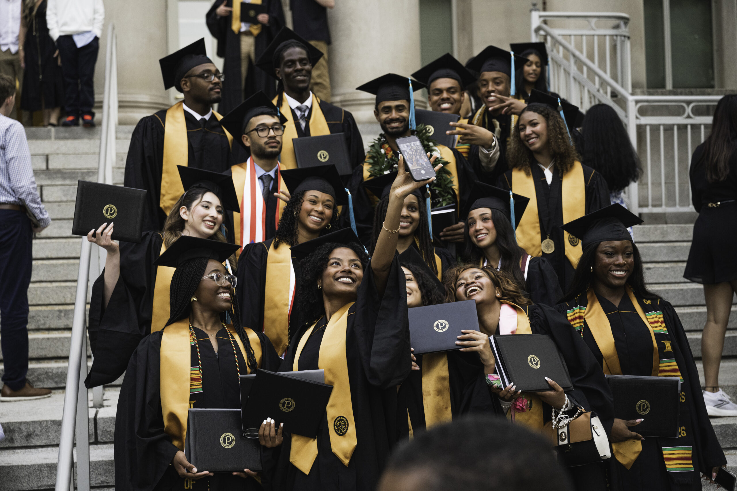 A group of graduates in black caps and gowns gather on campus steps, smiling and taking a selfie while holding diploma covers.