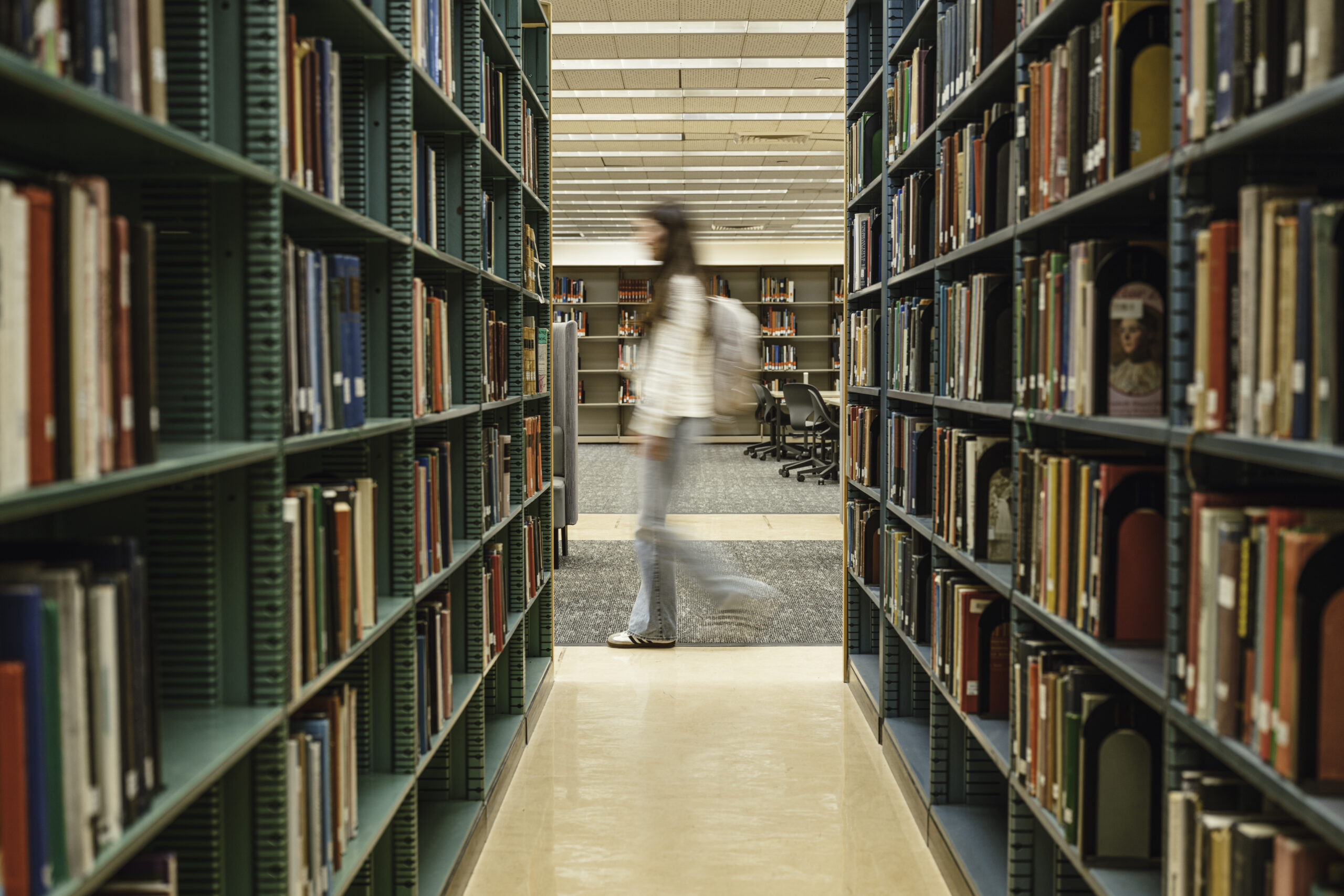 A blurred figure of a student with a backpack walks down an aisle between tall rows of green metal bookshelves filled with books in a library.