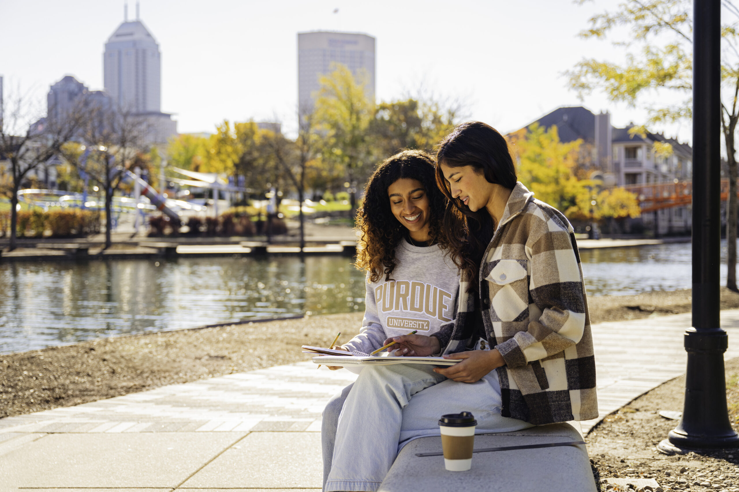 Two students sit together on a bench by a canal on a sunny day.