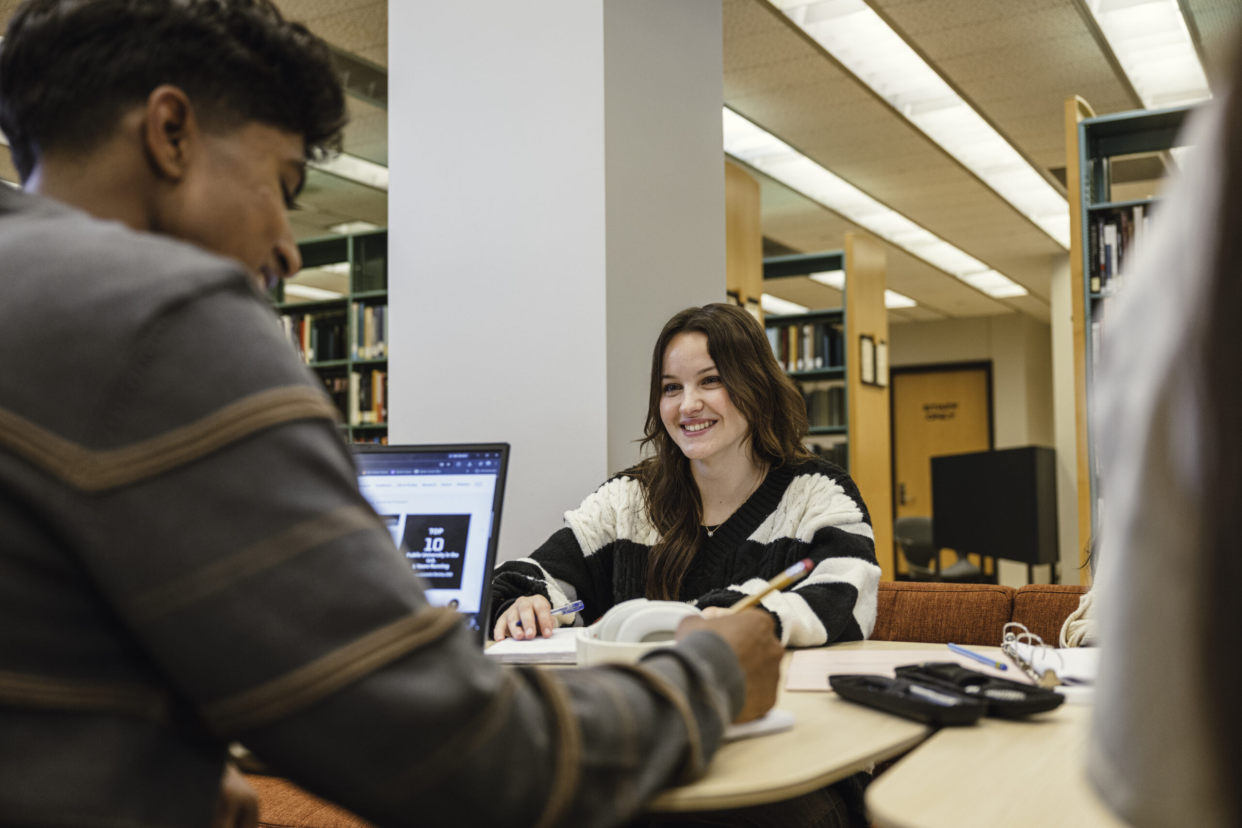 Two students study together at a desk inside a university library, with one student smiling toward their peer while working near a laptop and open notebooks.
